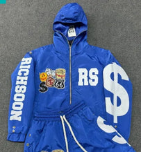 Load image into Gallery viewer, A bright blue hooded jacket and matching pants set laid flat on a gray textured surface. The jacket features a front zipper, large white 'RS' lettering on the chest, a large white dollar sign on the sleeve, and a vertical 'RICHSOON' print on the opposite sleeve.
