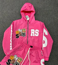 Load image into Gallery viewer, A bright pink hooded jacket and matching shorts set laid flat on a dark gray textured surface. The jacket has a front zipper, large white 'RS' lettering on the chest, white vertical lettering down one sleeve, and a decorative patch graphic on the left chest.